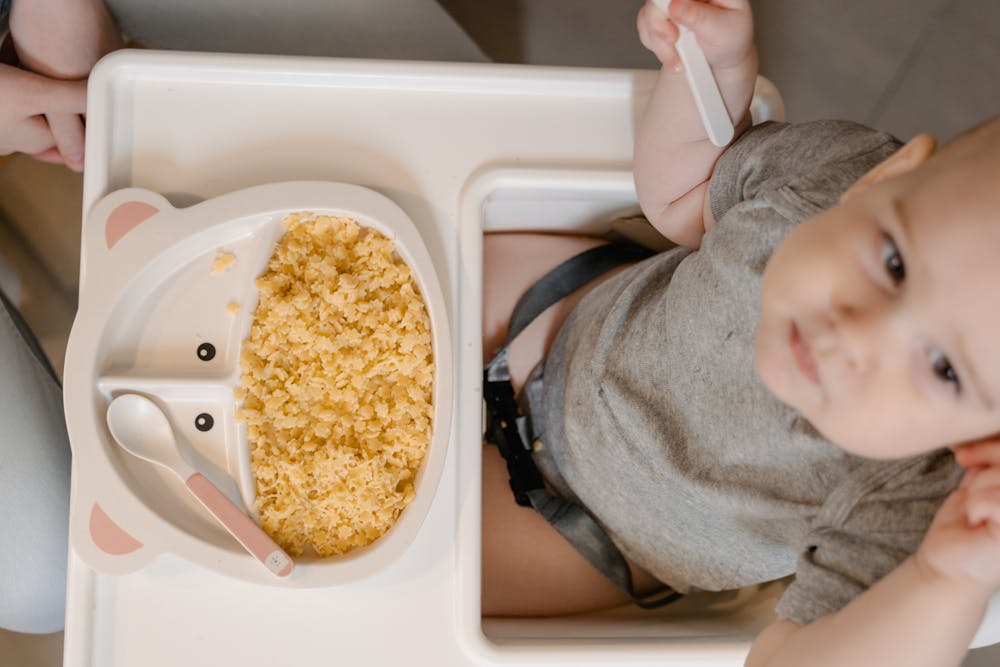 Baby sitting in a chair eating from a plastic plate and spoon.
